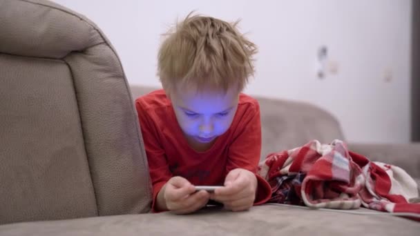 El niño está tumbado en el sofá con el teléfono inteligente en las manos y jugando al juego móvil o usando las redes sociales. El problema moderno de la dependencia de los chiquitines de los aparatos y el Internet desde la edad temprana — Vídeo de stock