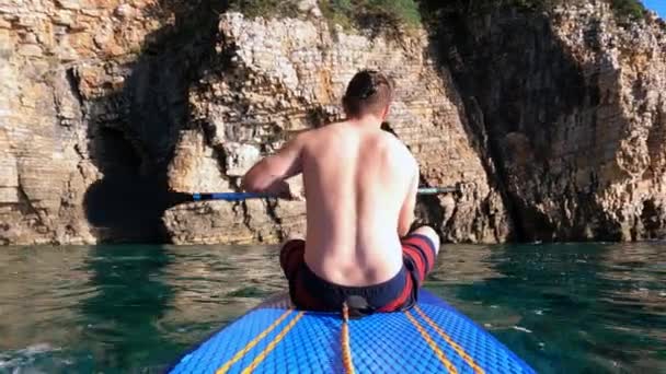 若い男行青いパドルボード海の上に古い急な崖に — ストック動画