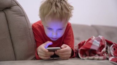 Gülümseyen şirin çocuk portresi kanepede uzanıyor ve telefondan oynanan bir oyunda neşeyle oynuyor. Gadget bağımlılığı. Gezici bağımlılık kavramı. İnternette sörf yapmak