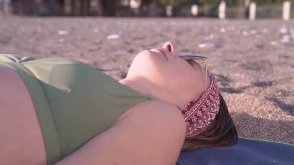 Młoda kobieta w okularach słonecznych opala się na plaży. Nieposłuszne dzieci lub zwierzęta bawią się zbyt aktywnie i uniemożliwiają kobiecie wygrzewanie się, przykrywając ją piaskiem. Szczotkuje ją irytująco. — Wideo stockowe