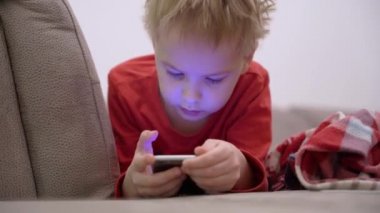 Küçük çocuk akıllı telefonuyla kanepede uzanıyor ve mobil oyun oynuyor ya da sosyal ağlar kullanıyor. Çocukların küçük yaştan itibaren cihazlara ve internete olan bağımlılığı sorunu. Mobil bağımlılık kavramı