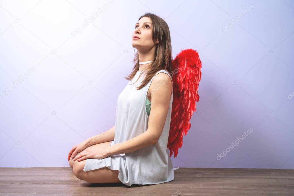 Attractive young woman in half-naked outfit with blood-red angel wings on back sits humbly on floor of empty room and looks up hopefully symbolizing opposing concepts of innocence and vice