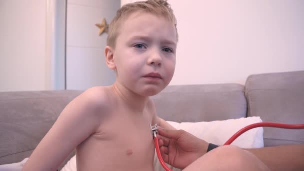 Врач осматривает расстроенного мальчика со стетоскопом на диване — стоковое видео