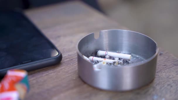 Woman ashes ciggarete into tray near smartphone at table — Stock Video