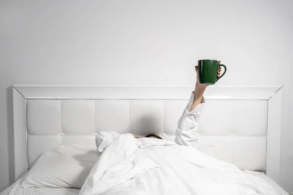 Woman holds green cup lying in comfortable bed with blanket — Stock Photo, Image
