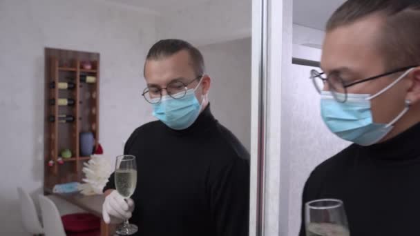 Giovane uomo in maschera medica protettiva celebra l'evento da solo a casa a causa di isolamento da un bicchiere di bevanda alcolica con il suo riflesso nello specchio. Nuova realtà durante la pandemia — Video Stock