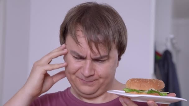 疑いの若い男は食事について考えるハンバーガーを食べたい — ストック動画