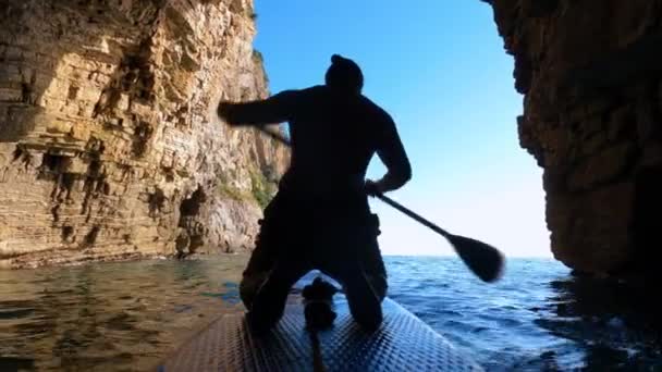 Silhouet van sporter roeiend SUP-board met peddel op zeewater door schaduwtunnel tussen oude steile kliffen achteraanzicht. — Stockvideo