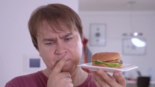 Thoughtful man holds delicious hamburger in kitchen closeup — Stock Video