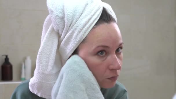 Mujer joven y agradable con toalla de baño envuelta alrededor de su cabeza después de la ducha limpia la cara de restos de cosméticos con un paño de limpieza de microfibra especial — Vídeo de stock