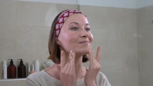 Жінка середнього віку наносить крем на обличчя, дивлячись у дзеркало — стокове відео