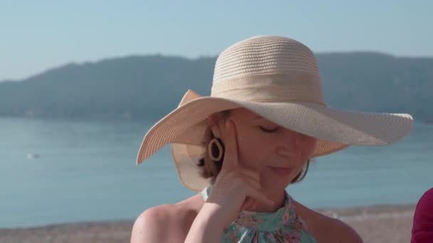 Porträt einer jungen Frau mit breitkrempigem Hut, die am Ufer des Meeres für die Kamera posiert, im Hintergrund verschwimmt der Berg. Schöne Dame auf Spaziergang — Stockvideo