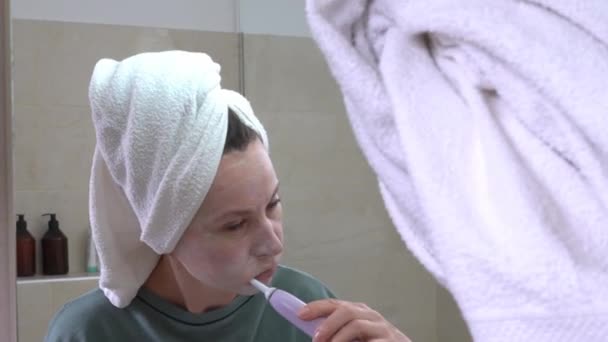 Junge süße Frau mit nährender Feuchtigkeitsmaske auf dem Gesicht und Handtuch um die Zähne gewickelt mit elektrischer Zahnbürste nach dem Duschen. Wellness-Wochenende — Stockvideo