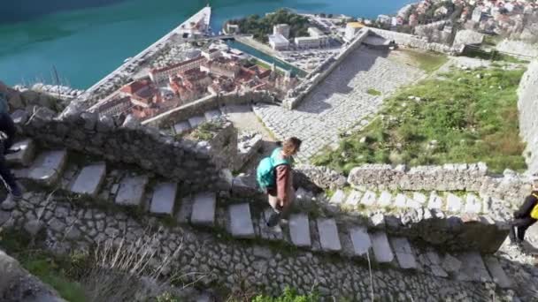 Toeristen met rugzakken dalen de oude stenen trap af van het verwoeste middeleeuwse kasteel naar de oude binnenstad aan zee. Reis naar historische plaatsen. Actieve levensstijl — Stockvideo