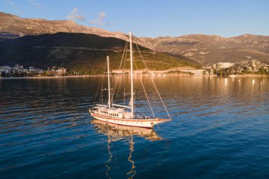 Büyük antika yelkenli yelkenli, güneşin altın ışıklarıyla aydınlatılarak, deniz koyunun ortasına demirlenir. Sakin su yüzeyi ve ormanla kaplı dağlarla kaplı güzel bir deniz manzarası.
