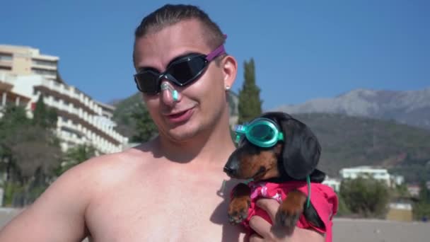 Jeune homme athlétique portant des lunettes de plongée et bouchon de nez pour la natation tient chiot teckel drôle en équipement de plongée et se expose devant la caméra avant d'aller dans l'eau, vue de face — Video