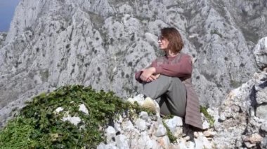 Güneş gözlüklü mutlu kadın, açık gri taşların üzerinde oturuyor.