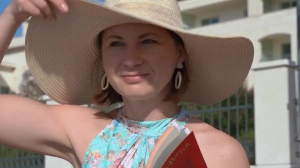 Красивая женщина в элегантной большой соломенной шляпе читает книгу на улице — стоковое видео
