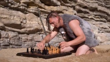 Kumlu plajda güneşli bir günde hayvan kürküne sarılı mağara adamı görüntüsüne bürünmüş Shaggy adam güvertede satranç taşları düzenler. Çağların karışımı