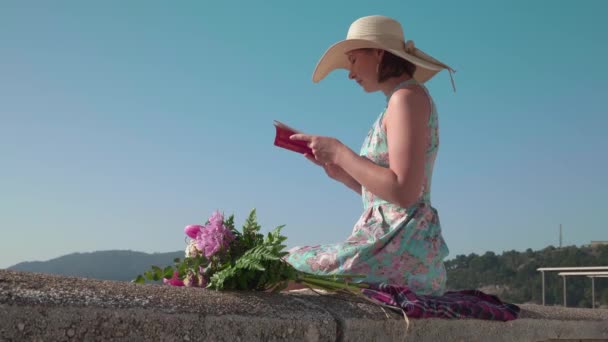Κομψή κυρία με ψάθινο καπέλο διαβάζει βιβλίο καθισμένος στο στηθαίο — Αρχείο Βίντεο