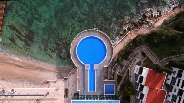 Drone se acerca lentamente a la piscina vacía abandonada en forma de ronda en el hotel resort en temporada baja, o parque acuático desierto. Playa de arena y fondo rocoso de hermoso mar claro, vista superior desde la altura — Vídeo de stock