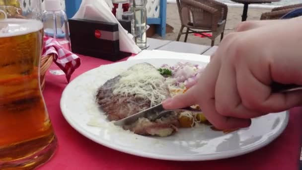 Το άτομο κόβει κομμάτι τηγανισμένου κιμά χρησιμοποιώντας πιρούνι και μαχαίρι στο καφέ, κοντά. Κρητικό ψητό πιάτο βαλκανικής κουζίνας - pljeskavica. Ωραίο γεύμα στο εστιατόριο με μια μπύρα — Αρχείο Βίντεο