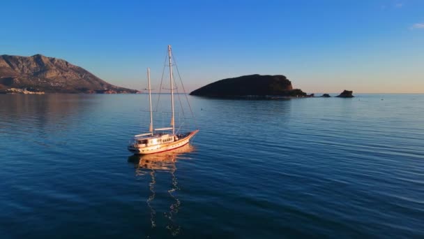 Drone com câmera voa após barco de pesca à deriva em águas calmas da baía perto de Budva, no Montenegro, cercado por montanhas e iluminado por raios dourados de nascer ou pôr do sol — Vídeo de Stock