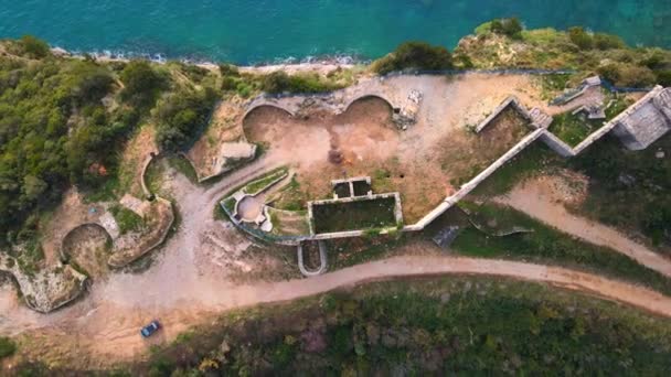 Drone με κάμερα υψώνεται πάνω από ερείπια του φρουρίου Mogren στη Μπούντβα του Μαυροβουνίου, που περιβάλλεται από δάσος και δρόμο, ο οποίος βρίσκεται στην κορυφή του βουνού που πλένεται από τη θάλασσα — Αρχείο Βίντεο