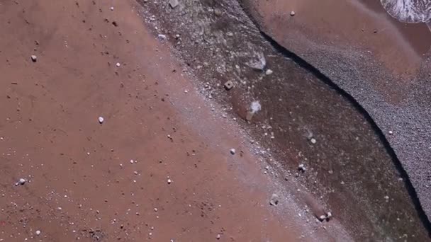 Mörk våt sand tvättas av skummande våg av turkos hav eller hav, bär småsten med sig i vatten, ovanifrån, drönare skytte från höjd. Någon hade lämnat fotspår på stranden längs strandlinjen. — Stockvideo