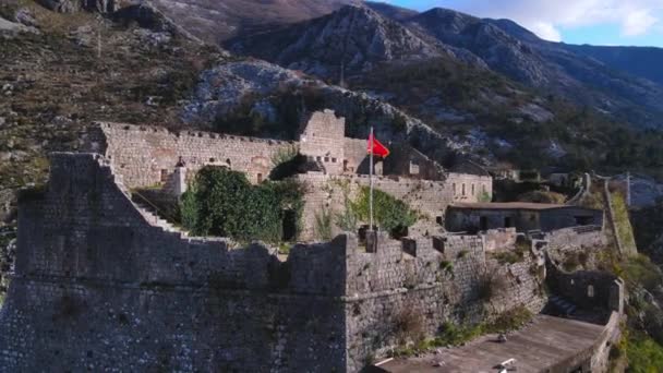 Drönare med kamera flyger runt medeltida österrikiska fästning ligger på toppen av berget i Kotor. Flagga Montenegro fladdrar i vinden, monterad på förfallen vägg — Stockvideo