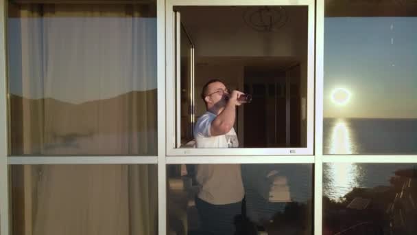 Glimlachende man drinkt bier staande bij hotel panoramisch raam — Stockvideo
