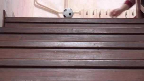 Osoba tlačí menší repliku fotbalového míče ze schodů, a hračka skáče po dřevěných schodech, zblízka, kamera se pohybuje za padajícím objektem, pohled zdola — Stock video