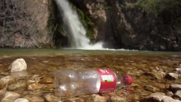 Botella de plástico con envoltura roja se encuentra en las rocas poco profundas del lago — Vídeo de stock