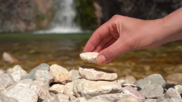 Person during a halt on the bank of mountain river on hiking trip tries to collect pyramid of stones, but everything falls apart and they have to start over, close up, waterfall in background — Stock Video