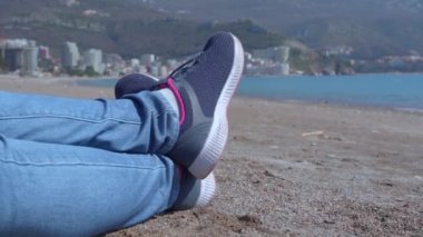 Kişi plajda uzanmış, güzel dağ manzarasına hayran kalıyor, dalgaları dinliyor, güneşli bir gün, çerçevede sadece ayakları spor ayakkabılarında, yakın çekim yapıyor.