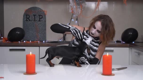 Joven mujer hosca con maquillaje de miedo en traje de esqueleto examina cachorro dachshund en la mesa, apartamento está decorado para la fiesta de Halloween. Monstruo sediento de sangre espeluznante quiere comer perro — Vídeos de Stock