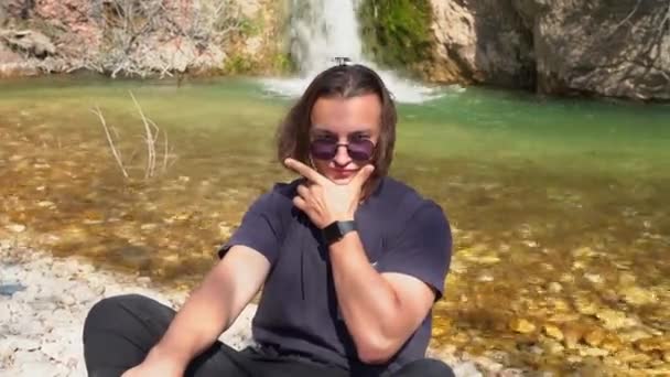 Güneş gözlüklü, uzun dağınık saçlı genç adam yürüyüş sırasında komik poz veriyor, küçük şelaleli bir dağ nehrinin kıyısında oturuyor. — Stok video