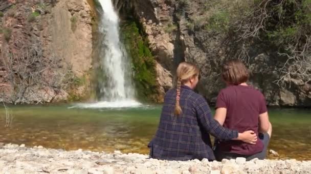 Dos jóvenes mujeres románticas se sientan en la orilla del río de montaña hablando entre sí, abrazando y admirando la cascada en una caminata, vista desde la parte posterior. Pareja viaja y lleva un estilo de vida activo — Vídeos de Stock