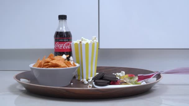 Set for pleasant movie viewing at home - bowl of nachos, box of popcorn, sweets and plastic bottle of Coca Cola without sugar. Food is on tray that someone is dragging across floor — Vídeo de stock