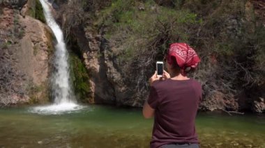 Kırmızı bandanalı turist kamp durağı sırasında şelalenin fotoğraflarını çekiyor, cep telefonu kullanıyor, arkadan görüşü var. Genç bayan blogcu seyahat vlog 'u için içerik oluşturuyor