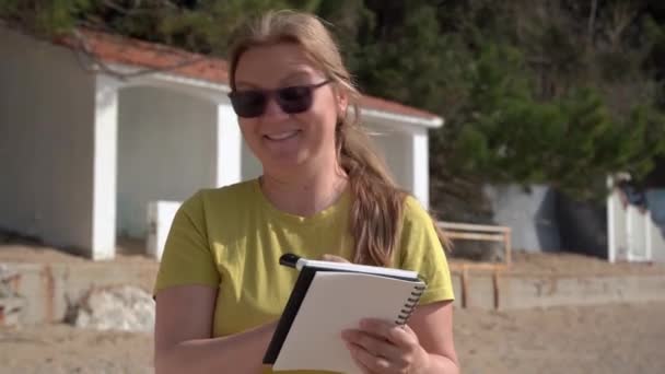 Positive Frau schreibt in Notizbuch, das am Strand steht — Stockvideo