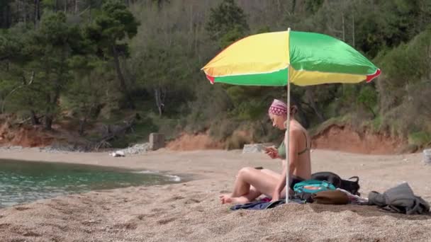 Kobieta w kostiumie kąpielowym siedzi pod parasolem plażowym ukrywając się przed palącym słońcem, aby się nie poparzyć i używając smartfona do pracy lub komunikacji. Dachshund pies przychodzi do osoby do zabawy z nią — Wideo stockowe