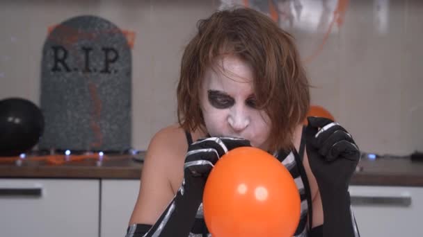 Disheveled young woman with creepy makeup in festive skeleton costume is getting ready for Halloween party so she inflates balloon to decorate apartment — Stock Video