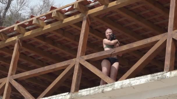 Junge Frau mit Kopftuch und zweiteiligem Badeanzug steht auf der Terrasse mit Holzgeländer, umarmt niedlichen Dackelwelpen und blickt von unten nach oben — Stockvideo