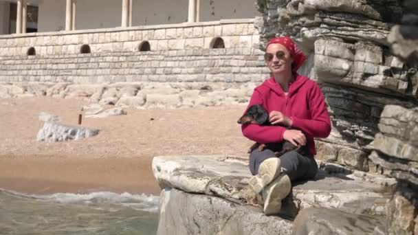 Jonge vrouw in bandana en zonnebril zit op rots met schattige teckel hond in haar armen, en bewondert prachtige zeegezicht, rust en mediteert tijdens de wandeling — Stockvideo