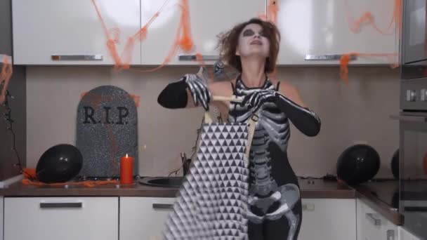 Jovem mulher vestindo traje de esqueleto com penteado desgrenhado e maquiagem temática estranha coloca em exposição na cozinha com dança em preparação para a festa de Halloween — Vídeo de Stock