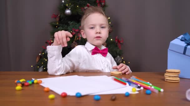 Enfant a reçu beaucoup de biscuits et de bonbons comme cadeau de Noël. Garçon est assis à table avec du papier et des crayons de couleur, manger des bonbons et penser à la lettre au Père Noël — Video