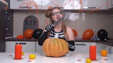 Şeytani iskelet kostümlü genç bir kadın Cadılar Bayramı partisi için balkabağı oyuyor. Kaşlarıyla şakacı jestler yapıyor ve bıçağını yalıyor.