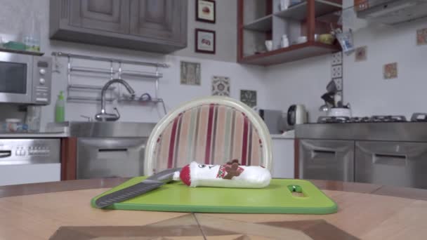 Lindo perro salchicha se sienta en la mesa en la que hay un cuchillo y un juguete suave en forma de muñeco de nieve, en la tabla de cortar de plástico. Mascota finge que va a cocinar en la cocina — Vídeos de Stock