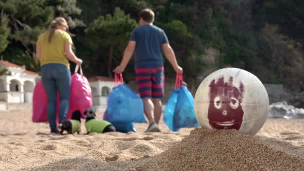 Young man and woman carry away packages, and dachshund dogs run after them. Volunteers have removed garbage on beach and are going to send it to recycling center. Ball with painted face rolled away — 비디오